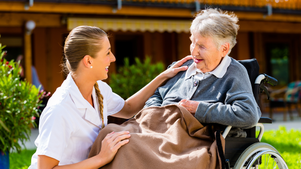 Elderly lady in wheelchair with a blanket over her legs, a care worker is kneeling next to her smiling with her hand on her shoulder. They are outside